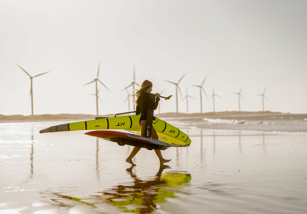 Instructora caminando en la playa de Icaraizinho con una tabla de wingfoil, aerogeneradores al fondo