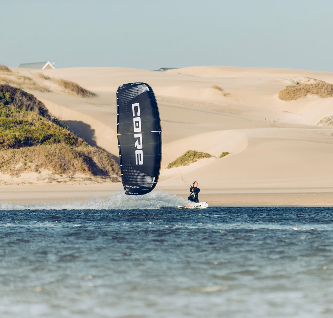 Rider flying a black Core kite between sand dunes, house in the background